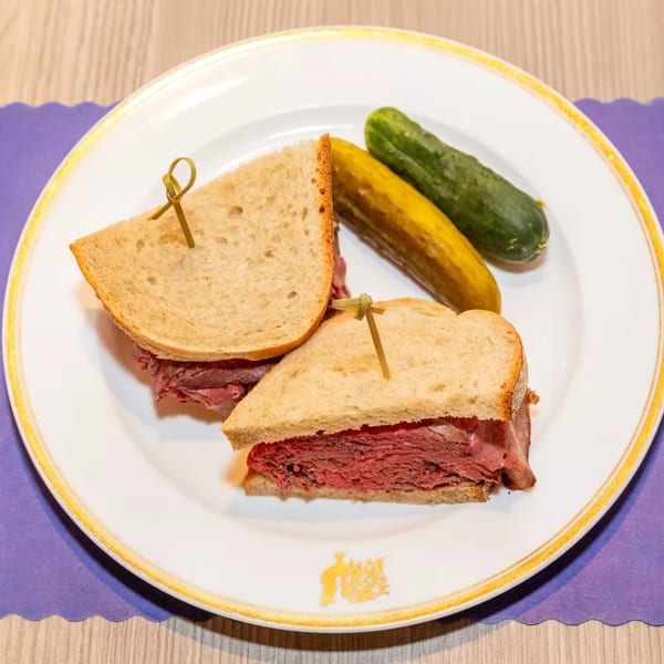 A pastrami sandwich on rye bread with two pickles on a white plate with gold trim.