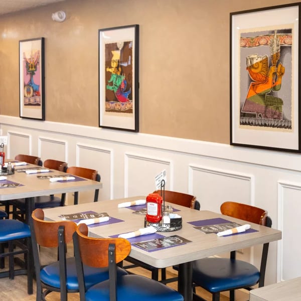 Restaurant dining room with light brown walls, white wainscoting, and tables set with blue placemats and menus.