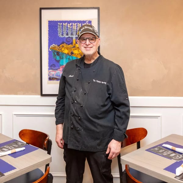 Smiling chef from The Pickled Herring deli in a black uniform and cap, standing by tables.