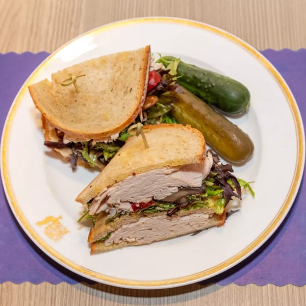 A turkey sandwich with lettuce, tomato, and mushrooms, served with pickles on a white plate.