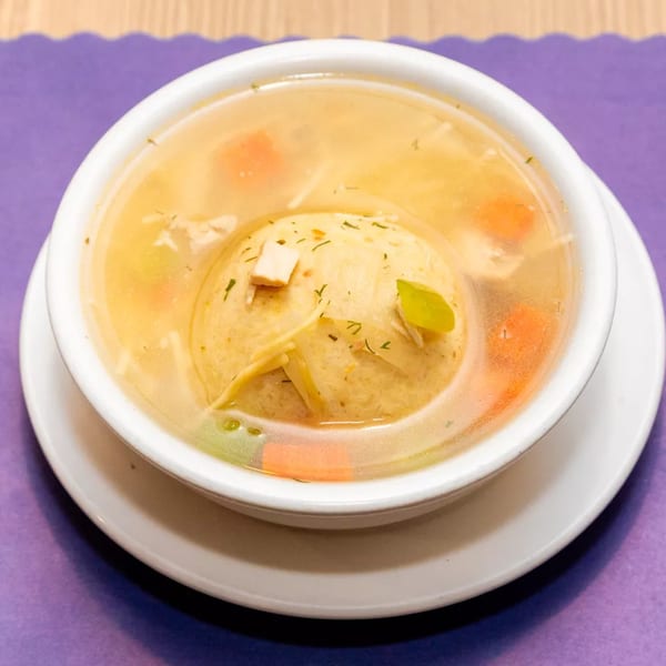 A bowl of matzo ball soup with carrots, celery, and noodles on a purple placemat.