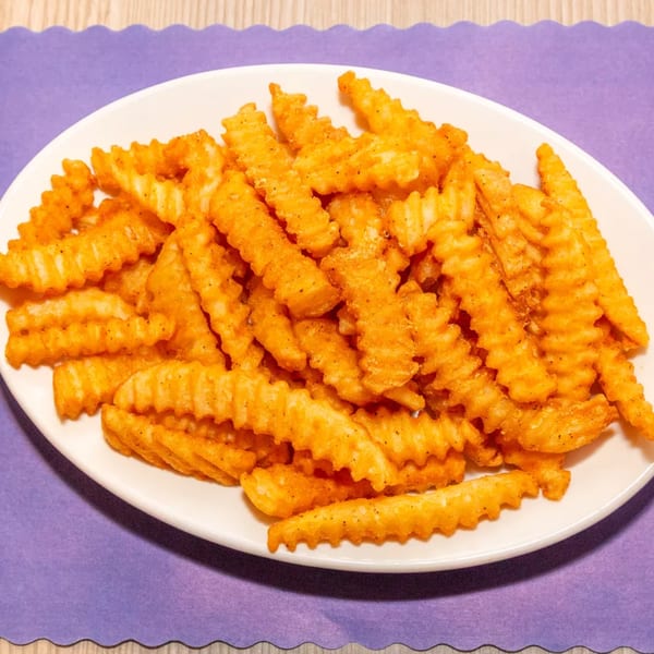 A white oval plate filled with golden, crinkle-cut fries, seasoned with pepper, on a purple placemat.