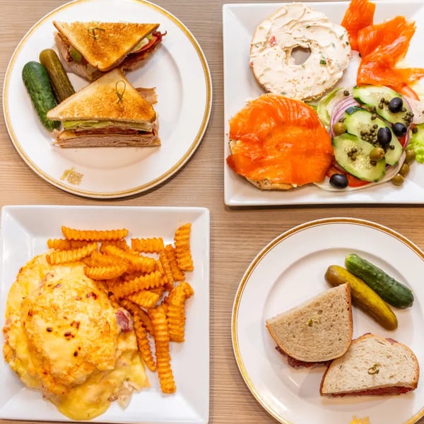 Four deli plates: club sandwich, lox and bagel, cheesy fries, and a pastrami sandwich with pickles.